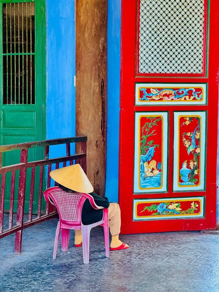 Femme au chapeau conique assise sur une chaise rose devant une porte rouge ornée, Vietnam